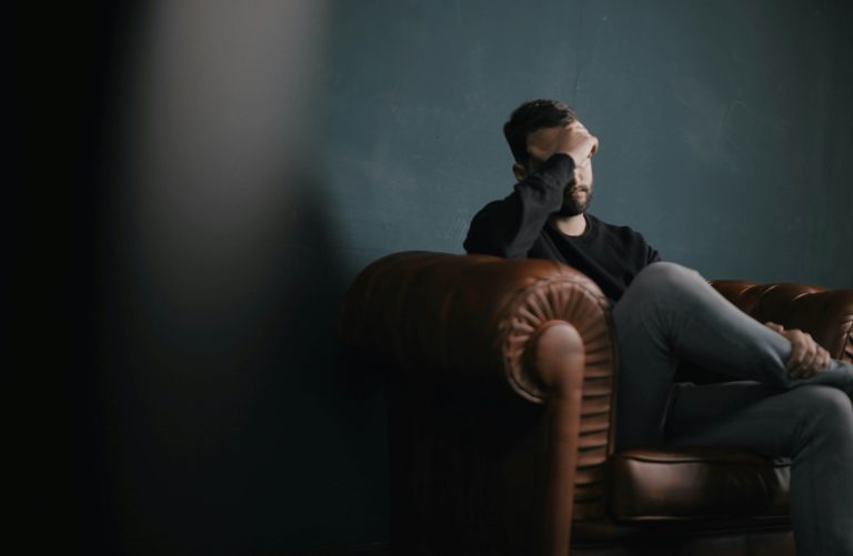 A man with dark hair dressed in a black shirt and jeans appears anxious and stressed due to high-functioning anxiety and sits alone on a brown leather couch with his head in hands.